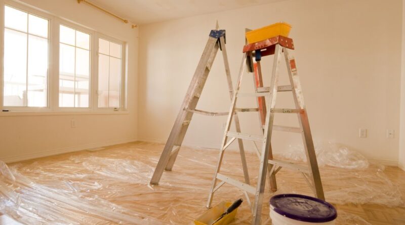 Ambiente em reforma de apartamento pequeno com escadas, balde de tinta e plásticos no chão, mostrando fase de pintura e preparo do espaço.